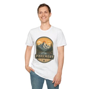 Peut inclure: Un t-shirt blanc à manches courtes avec un motif représentant un paysage de montagne, un soleil, des pins et une rivière. Le texte PINECREST NATIONAL PARK EST. 1932 est inscrit dans un emblème ovale à bordure sombre aux tons orange et bleu.