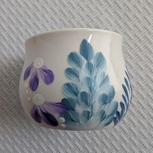 May include: A cream-colored ceramic mug with hand-painted floral designs. The mug features purple and blue flowers with white accents. The flowers are painted in a brushstroke style, giving the mug a unique, artistic look.