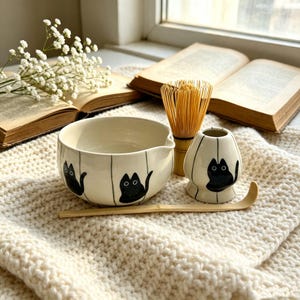 May include: A ceramic matcha bowl and a matching whisk holder, both featuring a cream-colored glaze with black cat illustrations and vertical lines. A bamboo whisk and a long, curved bamboo tea scoop are included in this set for tea preparation.