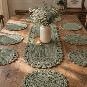 Puede incluir: Un juego de mantelería de ganchillo en color verde salvia que incluye un camino de mesa ovalado largo y siete manteles individuales circulares, cada uno con un borde festoneado decorativo. Un jarrón de cerámica blanca estriada con flores blancas.