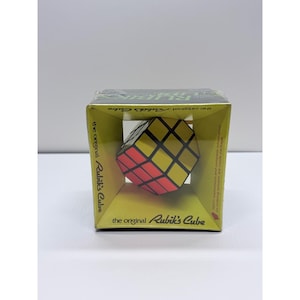 May include: A Rubik's Cube puzzle inside a yellow cardboard display box. The cube features a grid of small squares in red, yellow, and black. The packaging displays the text "the original Rubik's Cube" in a stylized black font on the front.