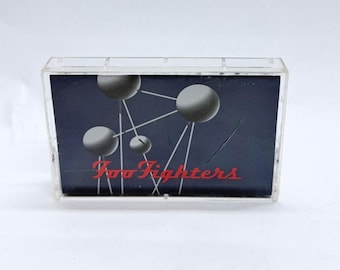 May include: A clear plastic cassette tape case containing an insert with a dark blue background, white lines connecting grey spheres, and the red text Foo Fighters printed across the lower portion of the graphic design on the front cover.