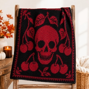 Puede incluir: Una manta rectangular de ganchillo con fondo negro y una calavera roja grande centrada en el medio. El diseño está rodeado de motivos de cerezas rojas con tallos y hojas, enmarcado por un borde decorativo rojo a lo largo de los lados.