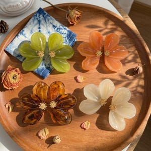 May include: Four pairs of flower-shaped hair clips in translucent green, orange, brown, and cream colors. Each clip features smooth, rounded petals and a small, textured gold-tone center. The clips are arranged on a round wooden surface.