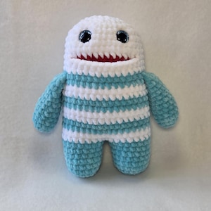 Puede incluir: Un peluche de ganchillo con forma de criatura, cabeza blanca, ojos oscuros y boca roja. El cuerpo presenta rayas horizontales de hilo azul claro y blanco alternadas. El juguete tiene dos brazos y dos piernas hechos de hilo suave y texturizado.
