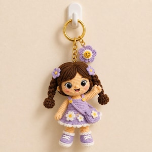Può includere: Un portachiavi a forma di bambola all'uncinetto con capelli castani intrecciati, che indossa un vestito color lavanda con applicazioni di margherite bianche e una borsa a tracolla abbinata. La bambola ha scarpe da ginnastica bianche e un ciondolo.