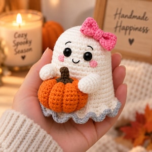May include: A small, white crocheted ghost figurine featuring a bright pink bow on its head, black bead eyes, and pink stitched cheeks. The ghost is holding a textured orange crocheted pumpkin with a dark brown stem and has a light gray scalloped edge.