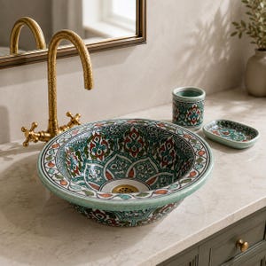 Könnte beinhalten: Ein rundes Aufsatzwaschbecken aus Keramik mit einem detaillierten Blumenmuster in Türkis, Rot und Weiß, ergänzt durch einen passenden Zahnputzbecher und eine Seifenschale. Dahinter befindet sich eine hohe, strukturierte goldfarbene Armatur.