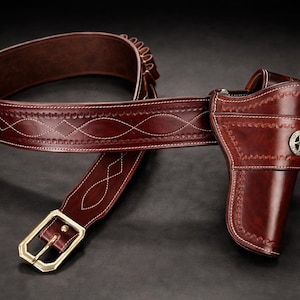 Pode incluir: Um coldre de couro castanho escuro para pistola preso a um cinto a condizer com fivela de latão. O cinto apresenta costuras brancas decorativas em padrão de diamante e presilhas integradas para munições. Um emblema de estrela prateada adorna o coldre.