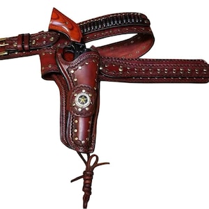 May include: A dark brown leather gun holster and matching belt set featuring intricate tooling, silver-toned metal studs, and a central silver star emblem. The holster contains a revolver with a polished wood grip and a row of ammunition loops.