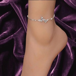 May include: A delicate silver-toned anklet featuring a central butterfly charm encrusted with clear, sparkling rhinestones. The chain is thin and metallic, designed to wrap around the ankle with a secure clasp, showcasing a bright, reflective finish.