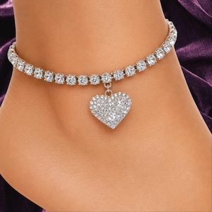 May include: A silver-toned anklet featuring a continuous row of clear, round-cut rhinestones. A heart-shaped pendant, fully encrusted with small, clear, sparkling rhinestones, hangs from the center of the chain.