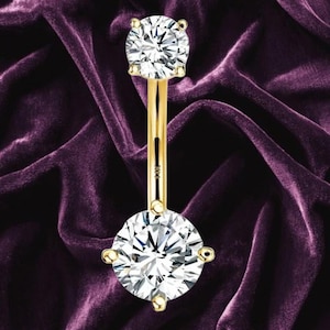 May include: A gold navel ring featuring a curved barbell with a smaller round clear gemstone at the top and a larger round clear gemstone at the bottom, each held in a four-prong setting. The metal bar is clearly stamped with the text 14K.