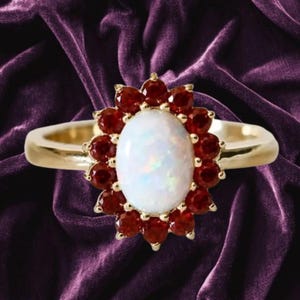 May include: A gold ring featuring a central oval-shaped white opal surrounded by a halo of twelve round-cut deep red garnet gemstones. The band is smooth and polished gold, creating a contrast with the vibrant red and iridescent white stones.