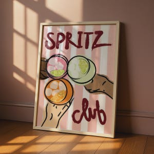 May include: A framed art print featuring a pink and white vertical striped background with the text SPRITZ club in dark red. The illustration shows three hands holding glasses filled with ice and citrus slices in orange, pink, and light green.