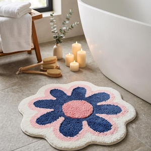 May include: A flower-shaped bath mat featuring a central pink circle surrounded by dark blue petals, all set against a white background. The mat has a textured, tufted surface and a scalloped edge, creating a distinct floral silhouette on the floor.