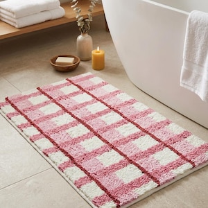 May include: A rectangular bath mat featuring a textured, tufted checkered pattern in shades of light pink, white, and dark red. The rug has a plush, shaggy pile construction with a grid design composed of alternating colored squares and thin red lines.