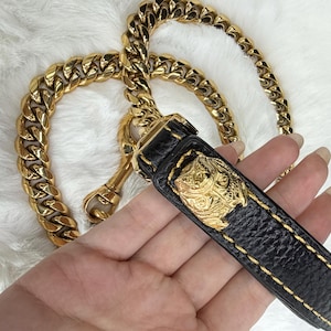 May include: A black leather leash handle with yellow stitching and a gold-toned metal lion head emblem, attached to a thick, polished gold-toned curb chain leash. The hardware includes a gold-toned swivel clasp for secure attachment.