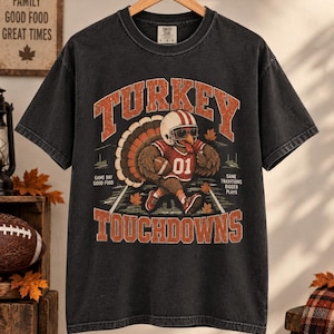 Op de afbeelding: Een antracietgrijs T-shirt met korte mouwen en een afbeelding van een kalkoen met een footballhelm, zonnebril en een shirt met nummer 01. De tekst luidt TURKEY TOUCHDOWNS, GAME DAY GOOD FOOD en SAME TRADITIONS BIGGER PLAYS.