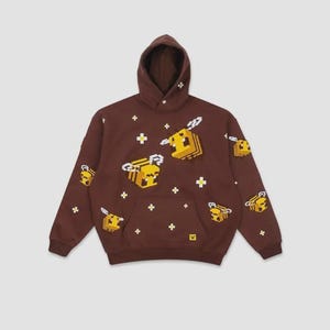 Peut inclure: Un sweat-shirt à capuche marron foncé orné d'un motif répétitif d'abeilles pixélisées en jaune, noir et blanc. Le design comprend de petites icônes de fleurs blanches pixélisées, une poche kangourou à l'avant et une petite étiquette logo jaune.