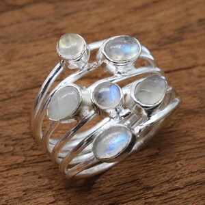 Puede incluir: Un anillo de plata de varias bandas con cinco cabujones de piedra lunar ovalados y pulidos. Las piedras presentan un aspecto blanco lechoso y translúcido con una sutil iridiscencia azul, engastadas en un diseño en espiral de plata pulida.