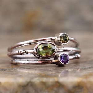 May include: A multi-band ring crafted from silver-toned metal featuring a textured finish. The ring is set with three distinct gemstones: a large central oval-cut olive green stone, a smaller round olive green stone, and a small round purple stone.