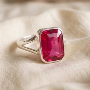 May include: A silver ring featuring a large, rectangular, emerald-cut vibrant pink gemstone set in a smooth metal bezel. The band of the ring has a split design that tapers as it connects to the central setting, showcasing a polished finish.