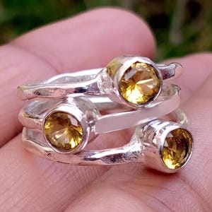 Peut inclure: Une pile de trois bagues en argent, chacune ornée d'une pierre jaune ronde à facettes sertie dans un chaton. Les anneaux en argent présentent une finition martelée texturée, créant un aspect superposé avec les trois pierres sous des angles variés.