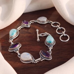 Op de afbeelding: Een zilveren schakelarmband met afwisselend druppel- en marquisevormige edelstenen, waaronder lichtblauwe larimar, dieppaarse amethist en iriserende maansteen. De armband is afgewerkt met een knevelsluiting en een ronde verlengketting.