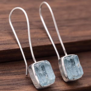 May include: A pair of drop earrings featuring rectangular, light blue, raw aquamarine stones set in polished silver bezels. Each stone is attached to a long, slender, curved silver ear wire that extends upwards from the top of the stone setting.