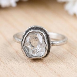 Peut inclure: Une bague en argent avec une pierre précieuse transparente, facettée et de forme irrégulière, sertie dans une monture texturée aux contours organiques. L'anneau présente une finition mate et martelée, donnant un aspect vieilli au métal.