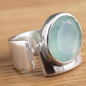 Puede incluir: Un anillo de plata con una gran piedra preciosa ovalada y facetada de color azul verdoso claro, montada en una banda de metal lisa y pulida. El anillo tiene un diseño abierto con una banda de plata ancha y reflectante que rodea la piedra.