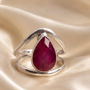 Op de afbeelding: Een zilveren ring met een druppelvormige, gefacetteerde donkerrode edelsteen in het midden. De ringband bestaat uit twee gladde, gepolijste zilveren draden die naar de edelsteen toe buigen, wat zorgt voor een open en gelaagd ontwerp.