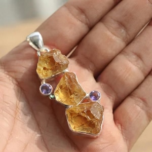 May include: A silver pendant featuring three raw, golden-yellow citrine stones arranged vertically, accented by two small, round, faceted purple amethyst stones set in silver bezels between the citrine pieces.