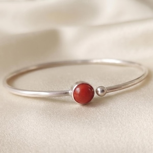 May include: A silver-toned open cuff bracelet featuring a smooth, round, reddish-orange stone set in a bezel on one end and a small, polished silver-toned sphere on the other end, creating a minimalist and balanced circular design.