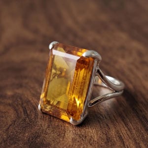 Könnte beinhalten: Ein Silberring mit einem großen, rechteckigen, facettierten goldgelben Citrin-Edelstein, der von vier Krappen gehalten wird. Die Ringschiene besteht aus zwei dünnen, parallelen Silberdrähten, die sich zur Basis hin vereinen.