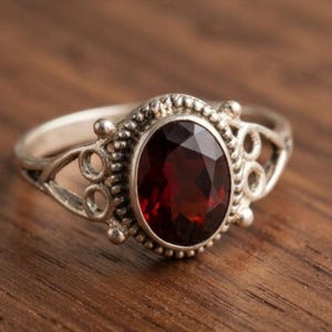 May include: A silver ring featuring a large, oval-cut, deep red gemstone set in a beaded bezel. The band displays an intricate openwork design with small circular accents on both sides of the central stone, creating a detailed metal frame.