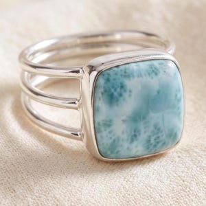 May include: A silver ring featuring a square, polished larimar gemstone with a light blue and white mottled pattern. The band consists of three parallel silver wires that connect to the bezel setting holding the stone in a smooth, square frame.