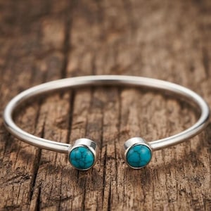 Könnte beinhalten: Ein silberfarbenes Armband in offener Form mit zwei runden, leuchtend türkisfarbenen Steinen mit dunkler Maserung an den Enden. Das Armband besteht aus einem glatten, polierten Metallband, das sich kreisförmig mit einer Öffnung vorne biegt.