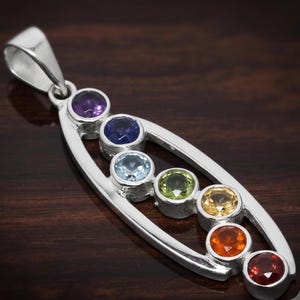 Puede incluir: Un colgante de plata con un marco ovalado alargado que contiene siete piedras preciosas redondas y facetadas dispuestas en una fila vertical. Las piedras muestran un espectro de colores que incluye violeta, azul, azul claro, verde, amarillo, naranja y rojo.