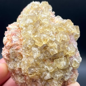 Peut inclure: Un amas de cristaux cubiques translucides jaune pâle formant un spécimen minéral dense et géométrique. La structure cristalline présente des bords nets et définis ainsi qu'une texture vitreuse, contrastée par une base de matière minérale rose clair.