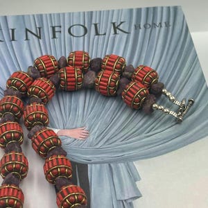 May include: A necklace featuring alternating cylindrical red beads encased in gold-toned metal cages and irregular dark purple stone beads. The piece is finished with a silver-toned toggle clasp consisting of a bar and a ring, creating a secure closure.