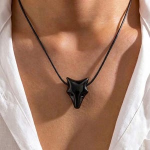 Pode incluir: Um pingente em forma de cabeça de raposa preta suspenso num colar de cordão preto fino. O pingente apresenta um acabamento liso e brilhante e um design angular estilizado que representa o rosto de uma raposa, centrado num cordão preto.