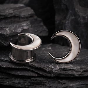 Puede incluir: Un par de expansores de oreja en forma de media luna con un marco de metal plateado pulido y una incrustación de concha blanca iridiscente. Un expansor se muestra de lado para revelar la forma cilíndrica, mientras el otro muestra la luna.