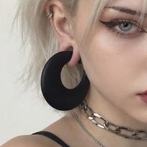 May include: A large, circular black ear gauge plug with a smooth, matte finish. The jewelry is worn in an earlobe piercing, featuring a wide, crescent-shaped design that creates a bold, solid black silhouette against the skin of the ear.