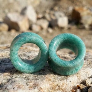 May include: A pair of circular ear plugs crafted from polished green amazonite stone. Each piece features a smooth, cylindrical shape with a hollow centre, showcasing the natural marbled texture and varying shades of teal and mint green throughout.