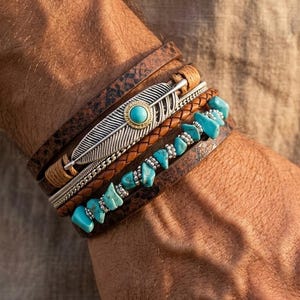 Könnte beinhalten: Ein mehrreihiges Armbandset bestehend aus einem breiten, strukturierten braunen Lederband, einer geflochtenen hellbraunen Lederschnur, einem silberfarbenen Federanhänger mit zentralem Türkisstein und einer Reihe aus unregelmäßigen Türkis-Splittern.