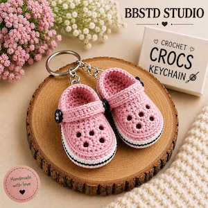 Puede incluir: Un par de llaveros de zuecos tejidos a ganchillo en color rosa con botones laterales negros y suelas blancas. Los llaveros están unidos a una anilla de metal plateado. Una caja blanca muestra el texto BBSTD STUDIO y CROCHET CROCS KEYCHAIN.