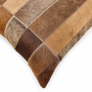 May include: A square throw pillow cover constructed from rectangular patches of genuine cowhide leather. The surface features a patchwork design with varying shades of brown, tan, and beige fur, joined together by visible light-colored stitching.