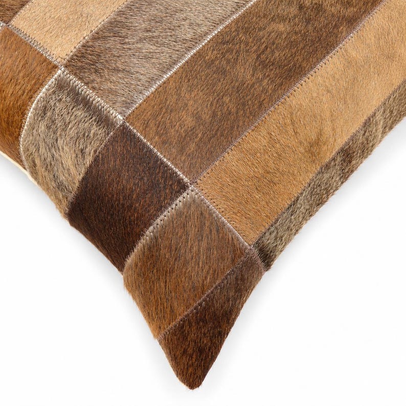 May include: A square throw pillow cover constructed from rectangular patches of genuine cowhide leather. The surface features a patchwork design with varying shades of brown, tan, and beige fur, joined together by visible light-colored stitching.